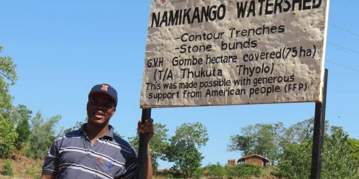 Man with Illini hat on standing next to a sign that says Namikango Watershed