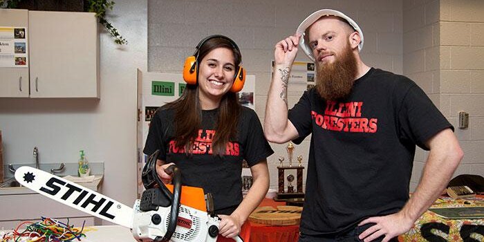 Two students with safety gear and a chainsaw